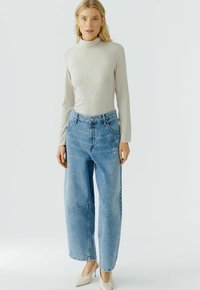 Lichtgrijze lange mouwen turtleneck gecombineerd met hoogsluitende, ruime lichtblauwe jeans. Model draagt beige puntige hakken. Effen achtergrond.