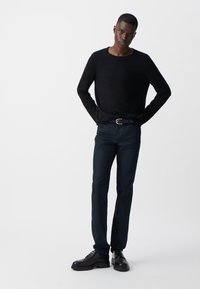 Maglione nero a maglia con maniche lunghe, abbinato a jeans slim blu scuro e scarpe nere con lacci. Modello in piedi con sicurezza, con le mani sui fianchi.