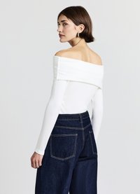 Weißes schulterfreies Rippstrickoberteil mit langen Ärmeln, kombiniert mit dunkelblauen, hoch taillierten, weitem Bein Jeans, die sichtbare Taschen und Nähte aufweisen.