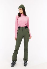 Vrouw in een groene beanie, roze langemouwen top, groene high-waisted skibroek en zwarte skischoenen op een witte achtergrond.