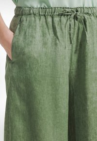 Zielone lniane spodnie z elastycznym pasem, sznurkiem do regulacji i bocznymi kieszeniami; tkanina o fakturze z lekko pogniecionym wykończeniem.