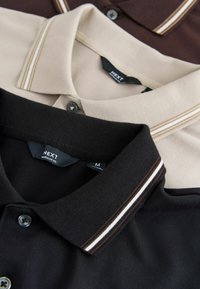 Trzy koszulki polo w kolorach czarnym, beżowym i brązowym, z paskami na kołnierzykach, guzikami oraz metkami z rozmiarem i marką "NEXT".