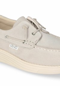 Scarpa in suede beige chiaro con punta arrotondata, cuciture decorative e occhielli in argento. Presenta una suola in gomma testurizzata e un'etichetta con logo.