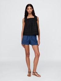 Top nero senza maniche con spalline arricciate e motivo floreale traforato, abbinato a pantaloncini in denim e sandali piatti. Sfondo grigio uniforme.