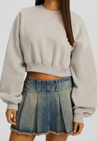 Kvinna klädd i en beige kort sweatshirt med ballongsärmar och en plisserad blekt blå denimminikjol med fransad nederkant.