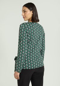 Langarmshirt in donkergroene stof met een geometrisch patroon in wit en lichtgroen. Voorzien van een strikdetail in de taille.