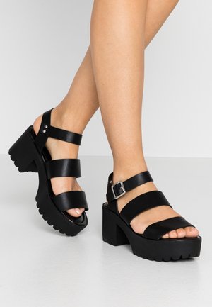 Sorte sandaler med chunky hæl, tre remme og en sølv spænde, båret på bare fødder mod en ensartet lys baggrund.