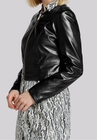 Chaqueta de cuero negra corta con un diseño ajustado, que presenta una cremallera central y una textura suave, combinada con un vestido de patrón blanco y negro.