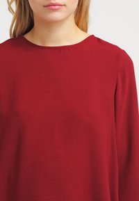 Top borgogna con scollatura rotonda, maniche lunghe e texture liscia. Design semplice senza motivi o decorazioni.