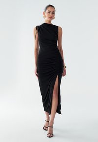 Sort draperet kjole med en skulderdesign, siden rynk, slids ved låret og glat tekstur. Inkluderer sorte slip-on sandaler.