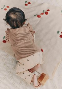 Suéter de punto marrón con mangas acampanadas, pantalones crema con patrones de cereza, bloque de madera sobre una alfombra texturada, bebé gateando sobre una superficie suave.