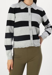 Harmaa ja musta raidallinen neuletakki, jossa on kauluksellinen pääntie, napitettava etuosa ja aaltoileva helma. Valmistettu pehmeästä neulemateriaaleista.