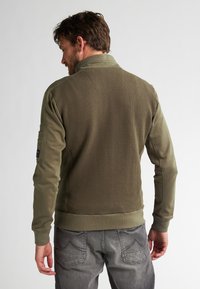 Olijfgroene jas met een gestructureerde achterkant, een gladde voorkant en geribbelde manchetten. Bevat een ritskraag en een opgestikte zak op de mouw.