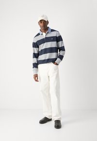 Sötétkék és szürke csíkos hosszú ujjú galléros póló, fehér nadrággal és fekete cipővel párosítva, gallérral és mellkason elhelyezett logóval.