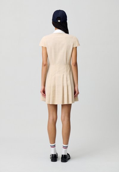Robe beige à manches courtes avec une jupe plissée, un col et un tissu texturé. Portée avec une casquette navy, des chaussettes blanches et des chaussures noires.