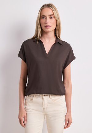 Femme portant un chemisier marron foncé à manches courtes en V, rentré dans un pantalon crème taille haute, debout devant un fond clair.