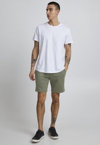 T-shirt en coton blanc, col rond, associé à un short en coton vert. Baskets noires, bras tatoués. Design simple, décontracté et sans fioritures.
