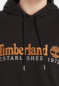 Felpa nera con cappuccio e logo arancione "Timberland", testo bianco "Established 1973" e emblema dell'albero ricamato sul petto, con stringhe pendenti.
