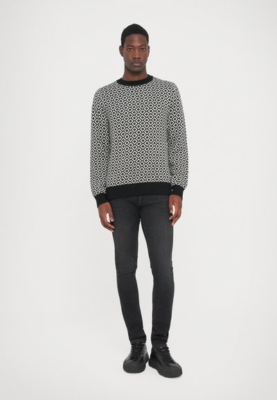 Black and white patterned sweater with diamond design, ribbed cuffs and hem. Worn with slim dark jeans and black shoes. Plain background.