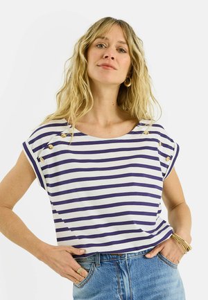 Mujer rubia con cabello ondulado que lleva pendientes de aro dorados, una camiseta de rayas blancas y azul marino con botones, jeans azules y pulseras y anillos dorados.