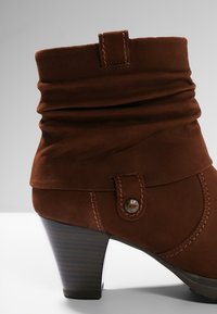 Bottine en daim marron avec un talon empilé de hauteur moyenne, une tige souple et une languette décorative cousue avec un bouton en métal.