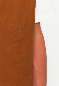 Gros plan sur une poche de gilet marron sur le torse d'une personne portant une chemise à manches courtes blanche, montrant la texture du tissu et les poils de bras.