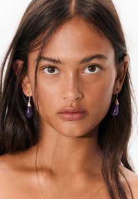 Lilla dråbeøreringe med gulddetaljer, med en glat tekstur og skinnende finish, båret af en model med langt mørkt hår.