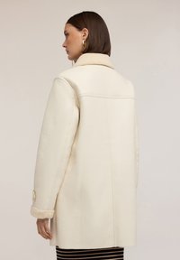 Cappotto crema in ecopelle con colletto in shearling, maniche lunghe e polsini con bottoni, caratterizzato da un taglio dritto e dettagli di cuciture minime.