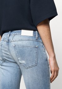 Vista trasera de unos jeans de mezclilla azul claro desgastados con una pequeña etiqueta blanca, combinados con una camiseta negra de manga corta sobre un fondo claro.