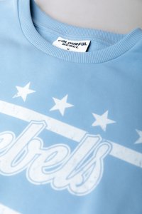 Lys blå sweatshirt med ribbestrikket hals, med et hvitt grafisk design som inkluderer ordet "rebels" og fem stjerner over.