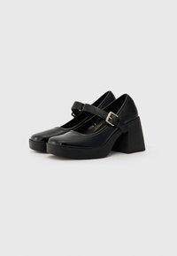 Black patent leather Mary Jane heels with a thick, block heel, rounded toe, and an adjustable strap featuring a silver buckle detail.