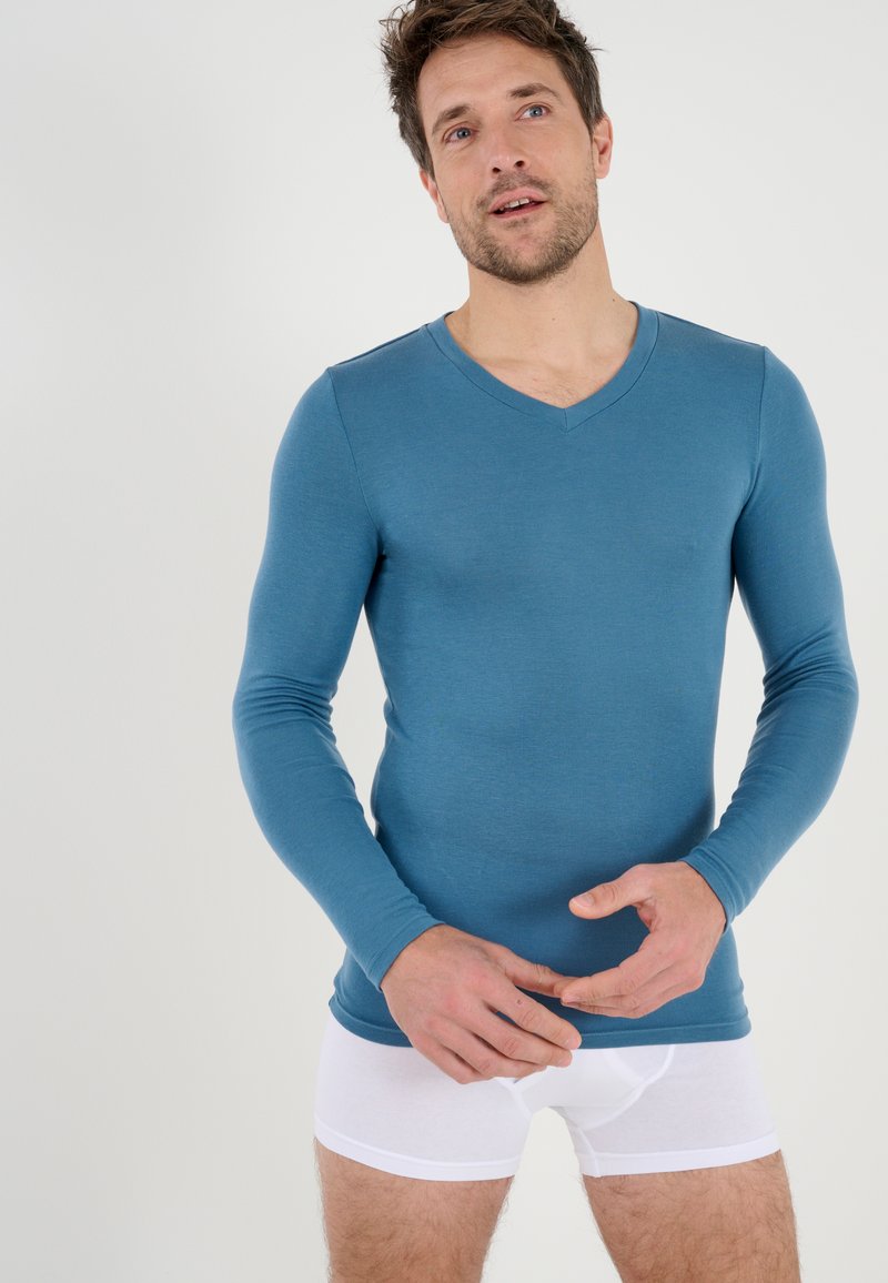 Top a maniche lunghe con scollatura a V in tessuto blu, abbinato a boxer bianchi. Texture liscia, design aderente che mette in risalto la forma del corpo.