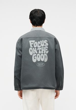 Person wearing a dark gray jacket with "Focus on the Good" and "Kaotiko Society" printed on the back, paired with light-colored pants.