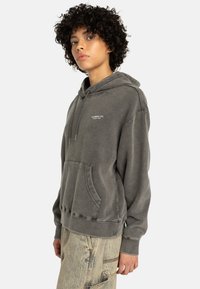 Szary hoodie z kapturem, z przednią kieszenią, o teksturowanym wykończeniu i wydrukowanym logo marki. Krótkie, szerokie rękawy oraz ribbowany dół.