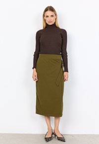Brauner gerippter Rollkragenpullover, olivgrüne Wickelröcke mit Bindegürtel und schwarz-weiße gestreifte High Heels. Glatte Stofftextur.