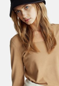 Beżowa bluzka z długim rękawem i kwadratowym dekoltem, o teksturowanej tkaninie i miękkim kroju, w zestawieniu z czarną kapeluszem typu bucket i naturalnie wyglądającymi włosami.