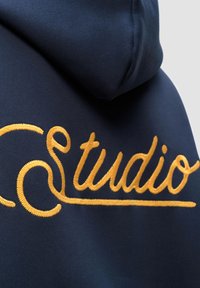 Felpa blu navy con grande testo ricamato "Studio" in filo giallo, realizzata in tessuto morbido e con una vestibilità rilassata.