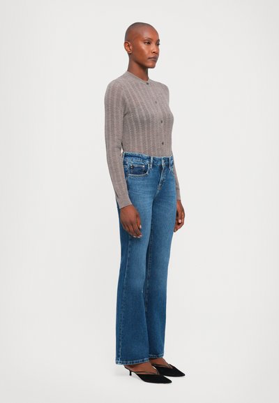 Cardigan grigio a coste con bottoni, abbinato a jeans a zampa d'elefante blu. L'outfit è completato da tacchi neri a punta.