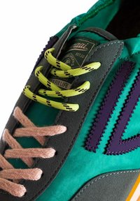 Scarpa sportiva con tomaia in tessuto verde, accenti in suede grigio, lacci a righe rosa e neri, e un logo viola testurizzato. Suola in gomma visibile.