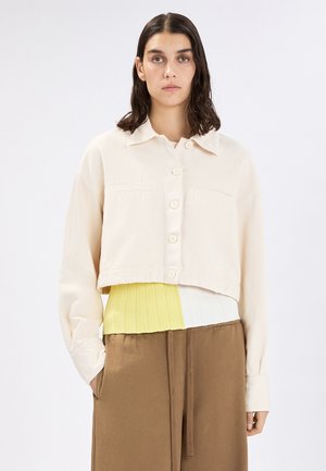 Donna con capelli scuri di lunghezza media che indossa una giacca crema corta con bottoni, un top giallo e bianco e pantaloni marroni con coulisse.