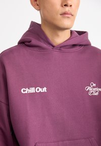 Lilla hettegenser i myk bomullsblanding, med hvit tekst "Chill Out" og "On Vacation Club." Snøring i hetten, ribbestrikkede mansjetter og kengurulomme.