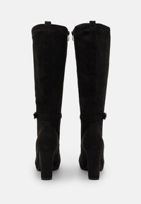 Botas de ante negro hasta la rodilla con una textura suave, tacones de bloque y cierre de cremallera. Presentan una correa decorativa con un pequeño detalle de hebilla.