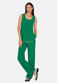 Groene mouwloze top met kantafwerking, gecombineerd met bijpassende losse broek. Gelaagd stof en glanzende zilveren schoenen maken de look compleet.