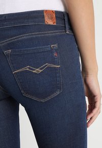 Mörkblå jeans har en slät textur, framträdande bakfickor med kontrasterande sömmar och en läderlogotyp på midjebandet.