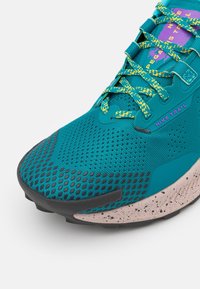 Nike trail løbesko med en turkis mesh-overdel, gule og blå snørebånd, sort ydersål og spottet beige mellemsål.