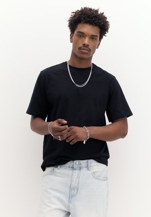 Mladý muž s kučeravými vlasmi, oblečený v čiernom tričku, svetlých džínsoch, so strieborným náhrdelníkom, náramkom a prsteňmi, stojaci na bielom pozadí.