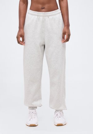 Άτομο που φορά ανοιχτό γκρι φόρμα και λευκά αθλητικά παπούτσια, όρθιο μπροστά σε απλό ανοιχτό φόντο, μεσαίο τμήμα και πόδια ορατά.