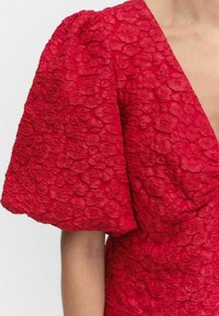 Czerwona sukienka z dużymi, teksturowanymi wzorami kwiatowymi i obszernym bufkami, ukazująca głęboki dekolt w serek i część ramienia noszącej.