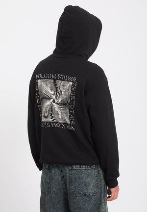 Personne portant un sweat à capuche noir avec un motif carré blanc déformé et un texte dans le dos, associé à un jean bleu délavé.