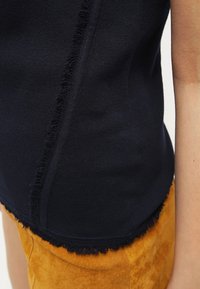 Top in blu navy a maglia con orlo frangiato testurizzato. Il tessuto è liscio, con un design aderente, mostrato su uno sfondo color senape.